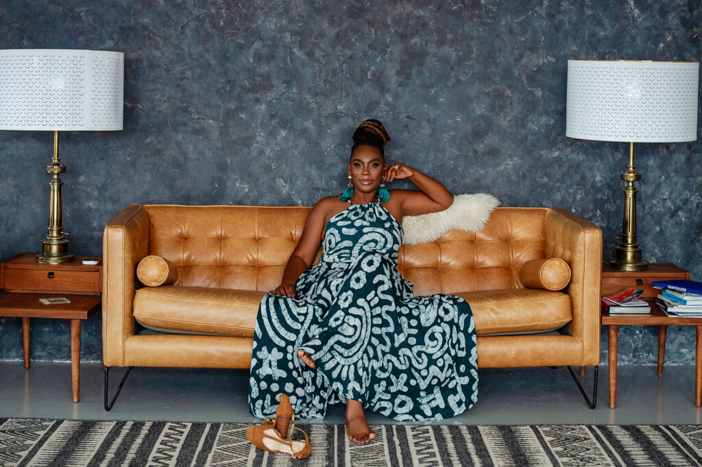Black woman in a flowered dress sitting on a tan leather couch smirking.