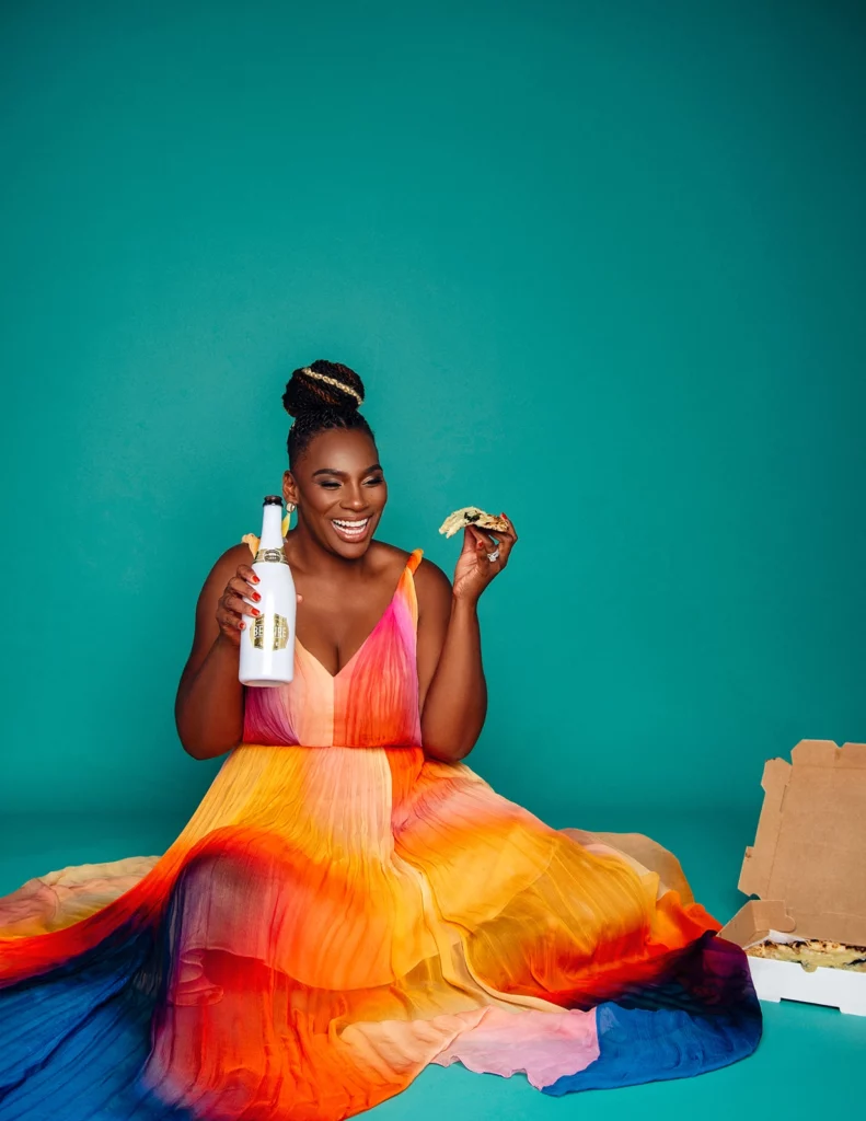 Black woman in multi colored dress sitting in the floor, eating pizza and drinking wine.